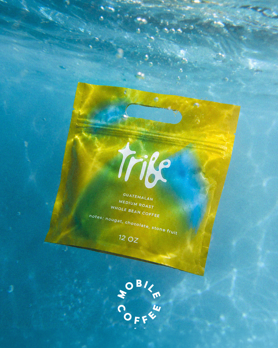 A bag of Tribe coffee floating underwater in a pool or ocean, with bubbles surrounding it. The bag is yellow and green with white text, indicating it is Guatemala medium roast whole bean coffee.