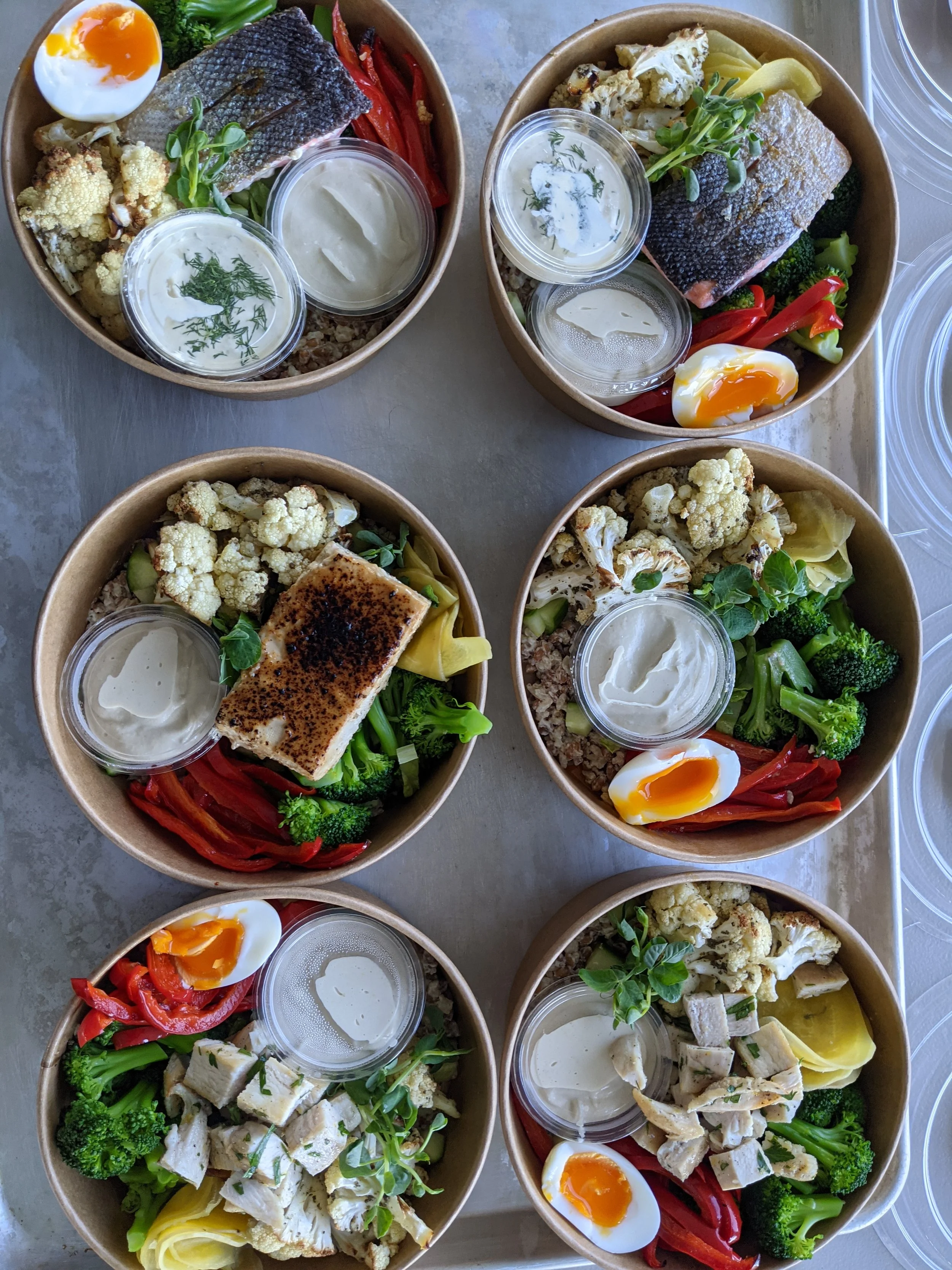 Assorted food bowls with ingredients like fish, chicken, roasted vegetables, quinoa, soft-boiled eggs, and sauce cups.