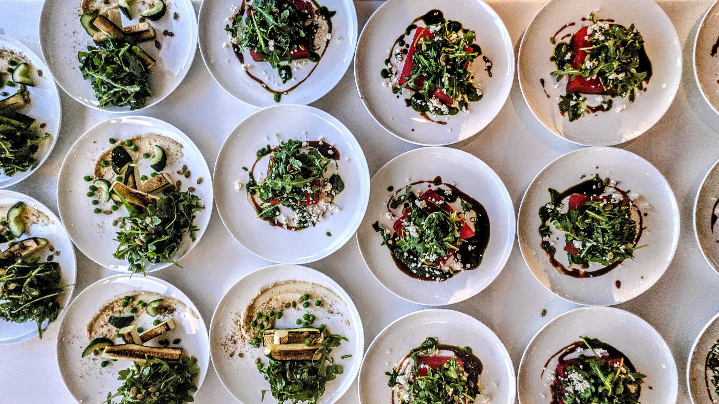Plates of gourmet salads with arugula, pieces of watermelon, cheese, and dressing arranged in rows.