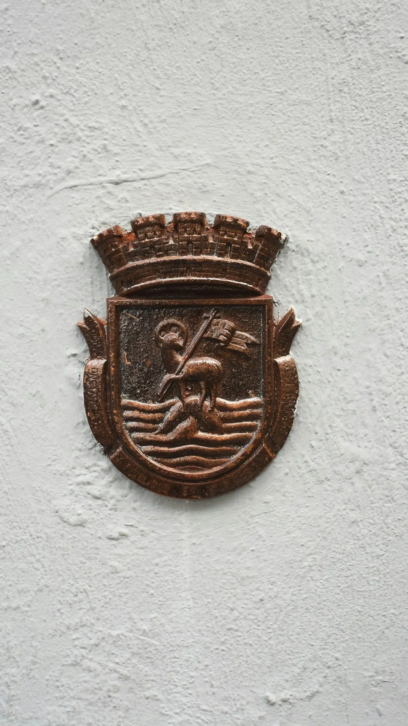 A bronze crest featuring a person walking through water, holding a flag, with a city wall or castle crenellation above, mounted on a light-colored wall.