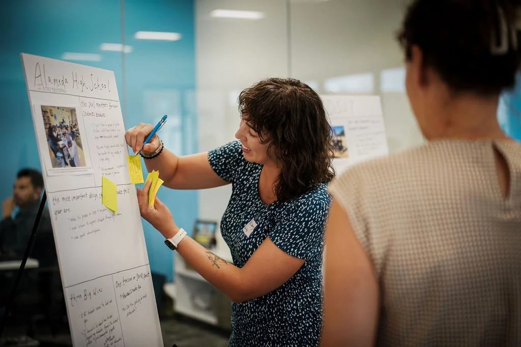 A woman presenting a project board titled "Alameda High School" at a classroom or conference setting, with an audience member in the foreground.