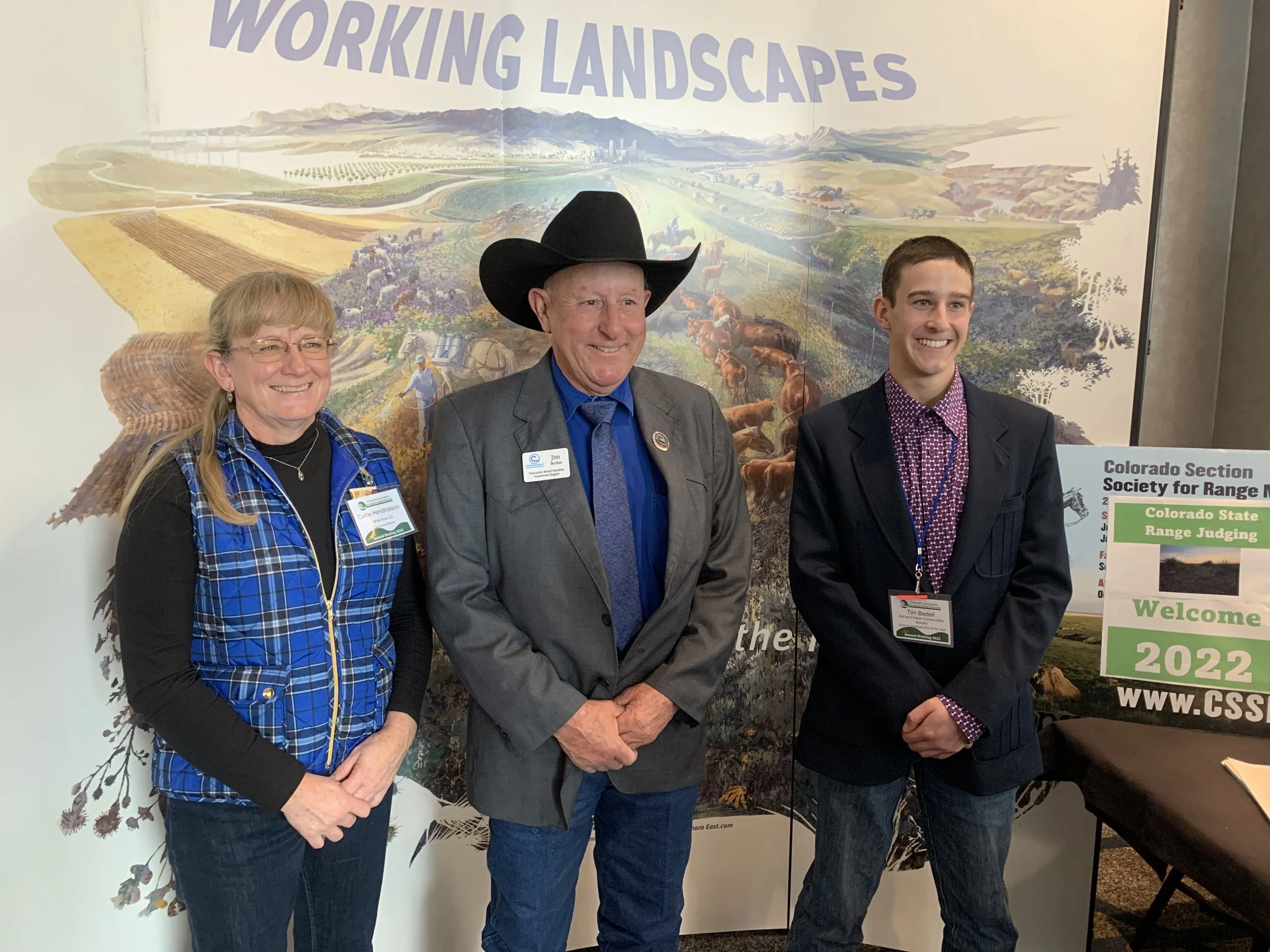 Three people standing in front of a large poster that says 'WORKING LANDSCAPES'. The woman on the left is wearing glasses, a black shirt, and a blue plaid vest, smiling. The man in the middle is wearing a black cowboy hat, gray blazer, and blue shirt, also smiling. The young man on the right is wearing a black blazer, patterned shirt, and is smiling. There is a sign on a table to the right that says 'Colorado Section Society for Range Management' and 'Welcome 2022'.