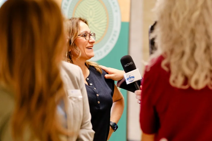 A woman being interviewed by a group of people, with a microphone in hand, in front of a green emblem.