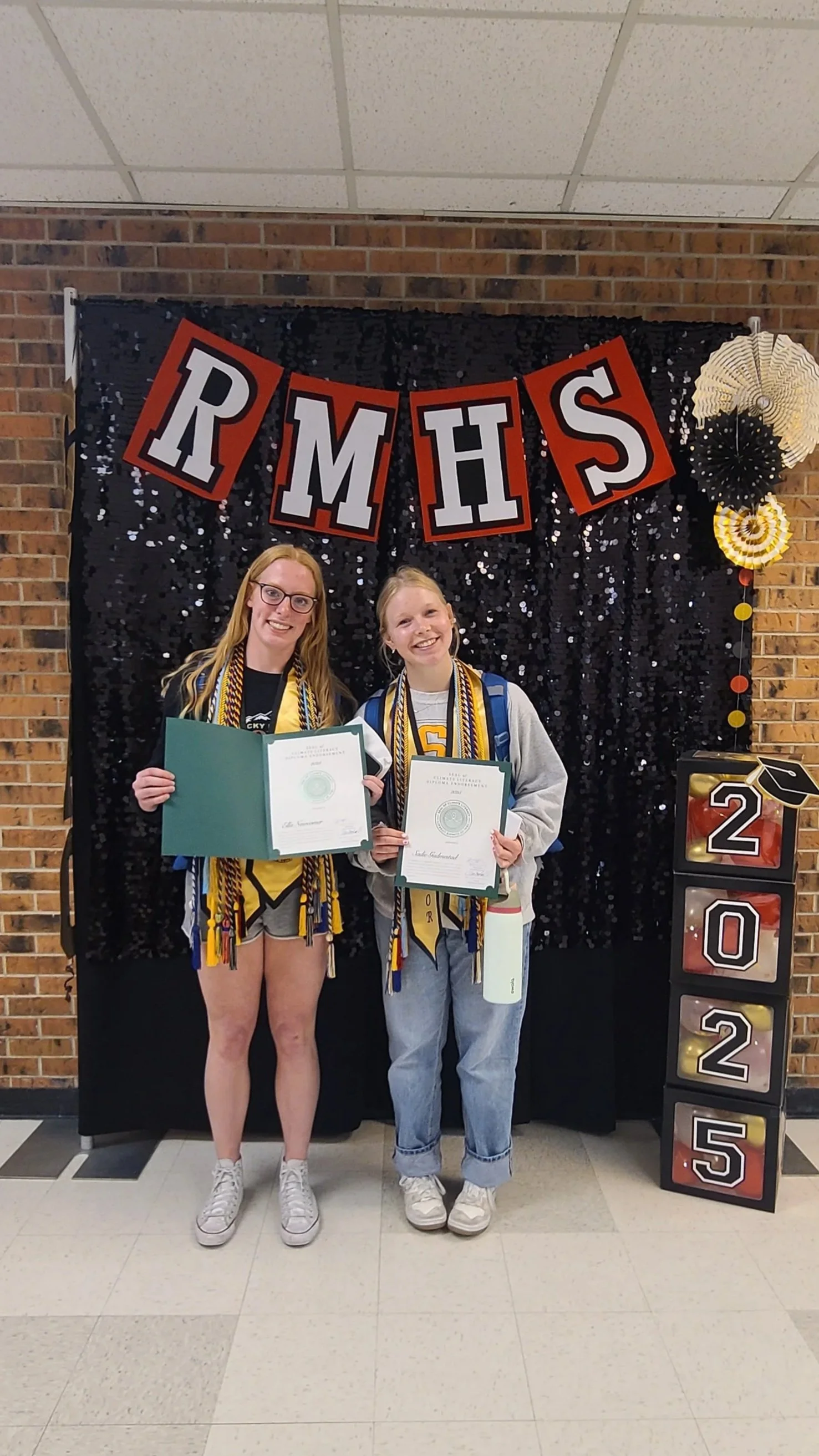 Two girls standing side by side, holding certificates, smiling for a graduation photo at RMHS, with a black sequin backdrop and decorations including a banner with the letters 'RMHS', the year '2025', and celebratory paper fans.