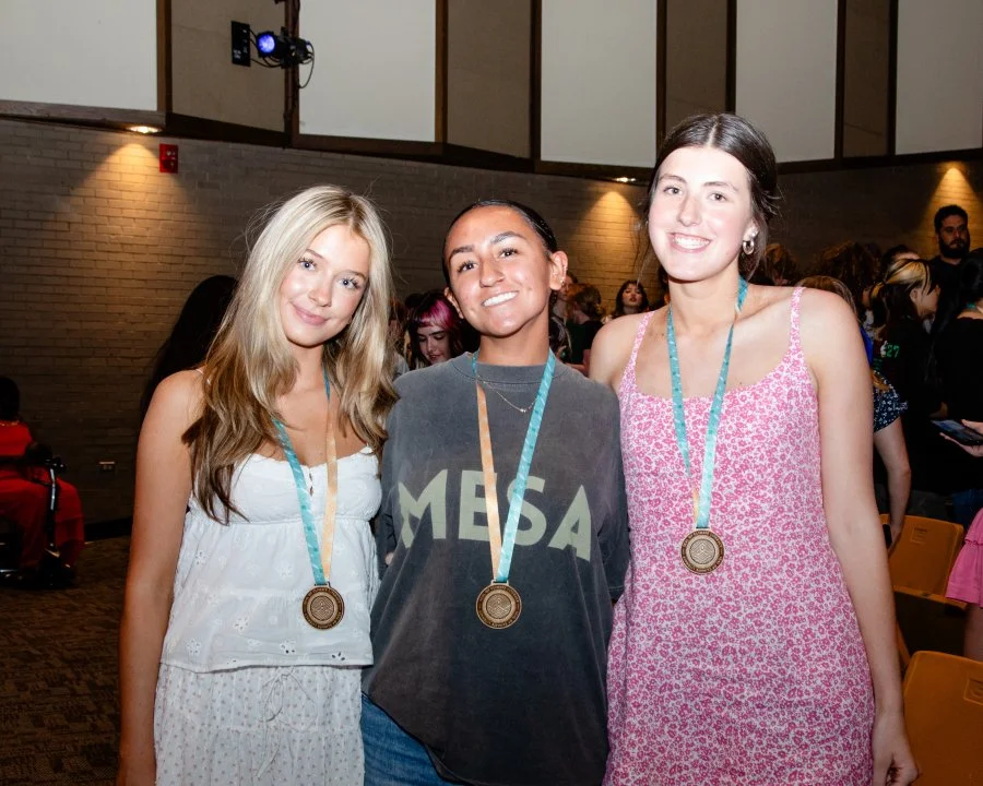 Three young women stand together, wearing medals around their necks, at an indoor event or ceremony. The woman in the middle wears a casual gray T-shirt with "MESA" printed on it, while the women on either side wear light dresses, one in white and the other in pink floral.