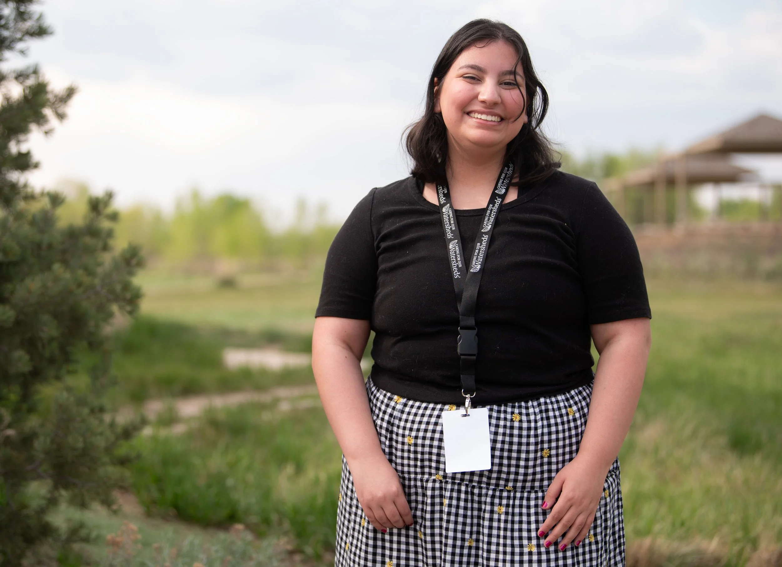 A woman standing outdoors during daytime, smiling, wearing a black shirt and a black and white checkered skirt with small yellow flowers, with a lanyard and badge around her neck, in a grassy area with trees and structures in the background.