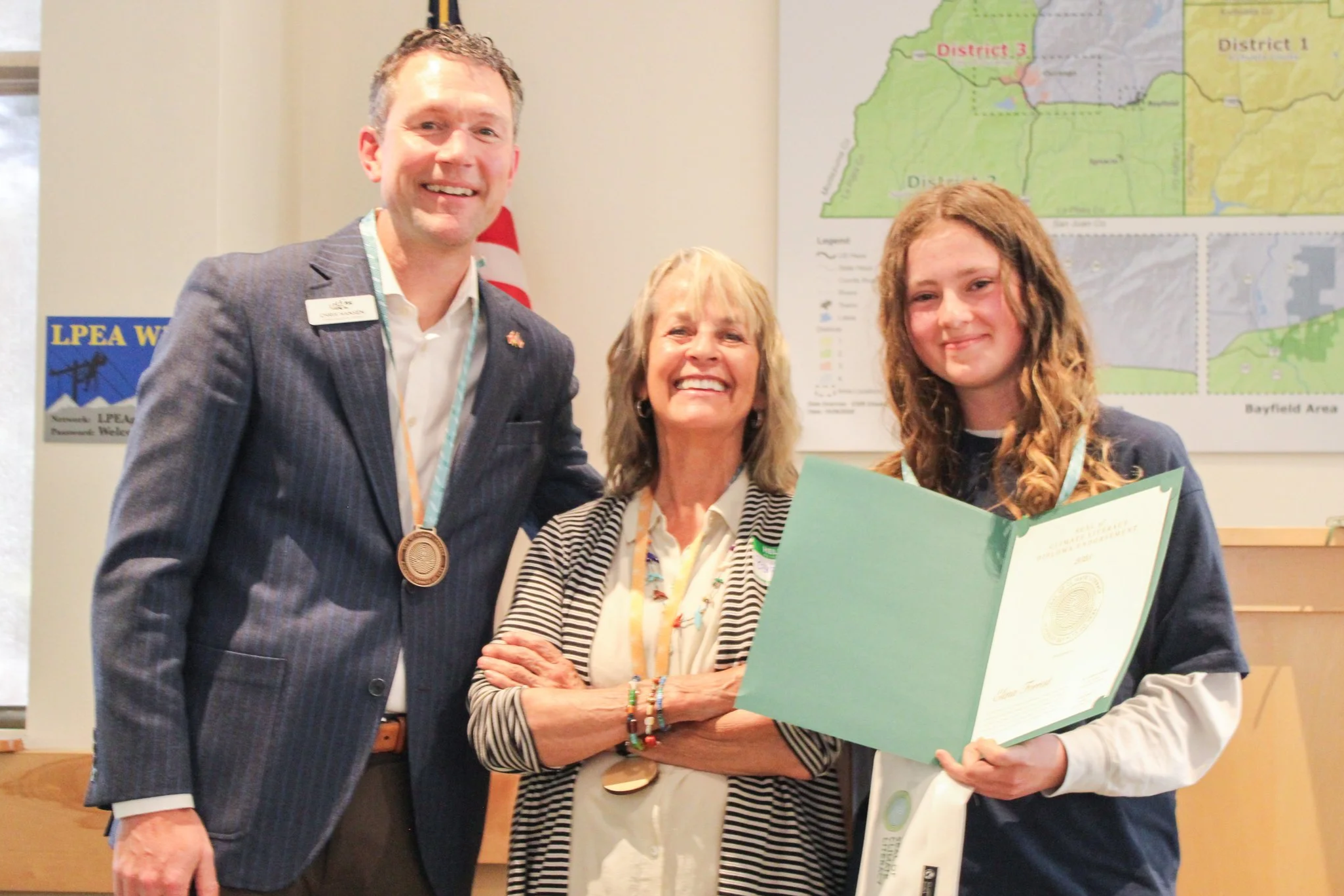 A young girl receiving an award certificate from a woman, with a man and a woman standing next to them, in a room with a map of geographic districts on the wall.