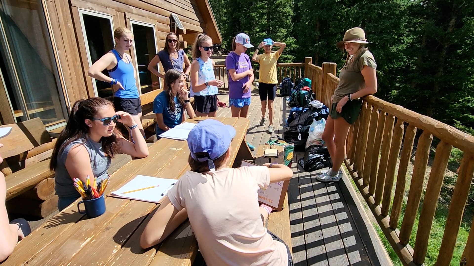A group of young girls and a woman are gathered on a wooden balcony surrounded by trees. Some girls are seated at a picnic table with notebooks and supplies, while others are standing and listening to the woman, who appears to be instructing or talking to them. The scene looks like an outdoor summer camp or educational activity.