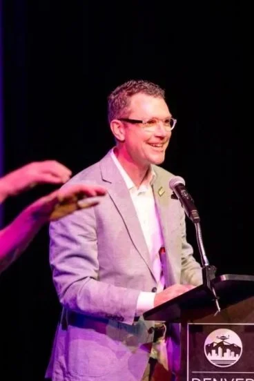 Man in gray blazer and glasses speaking at a podium with a microphone, with a hand in the foreground, against a black background.