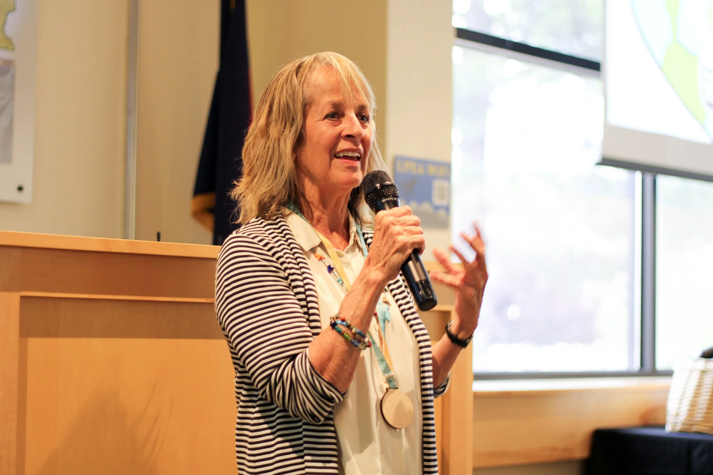 A woman with blonde hair wearing a striped blazer and a large circular necklace, speaking into a microphone at a presentation or conference, with a window in the background showing trees outside.