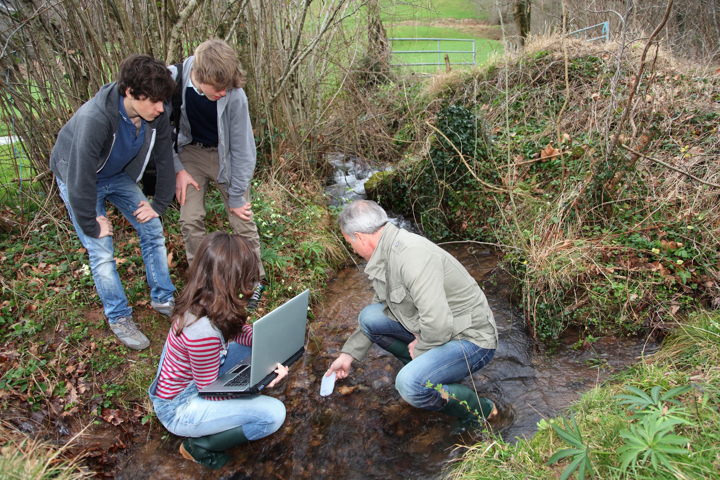 A group of five people, including children and an adult, gathered by a creek in a wooded area, examining the water with a laptop and a sample container.