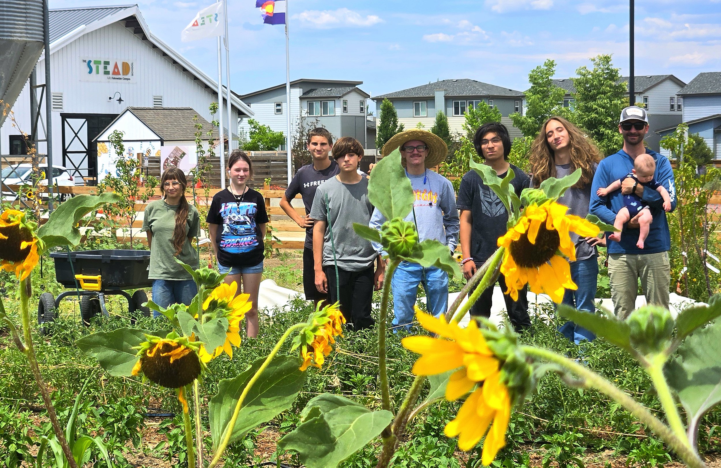 A group of nine people standing in a garden with sunflowers and other plants, with houses and a community center building in the background. The group includes children and adults, some smiling and dressed casually. One adult is wearing a wide-brimmed hat and holding a baby. The setting appears to be a sunny day in a suburban neighborhood.