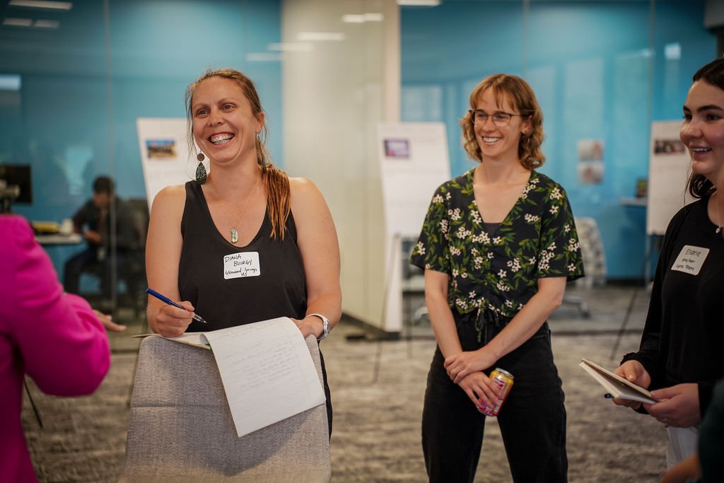Three women smiling and talking at a professional event or conference in a modern indoor space with blue walls and white display boards.