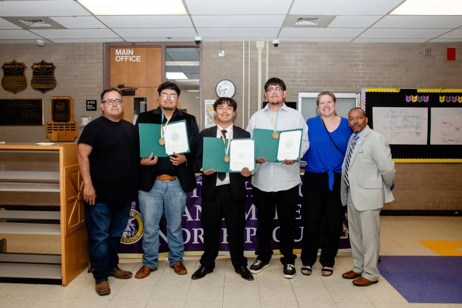 Group photo of five people, three young men holding awards and wearing medals, with two adults standing beside them, inside a school hallway with a sign that reads 'Main Office'.