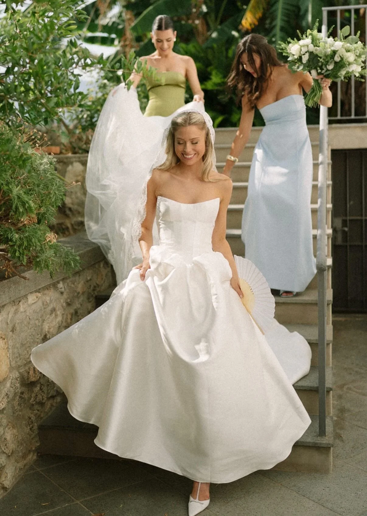 A woman in a white wedding dress with a veil, smiling as she descends outdoor stairs, with two women behind her helping with her dress. One woman holds a bouquet of white flowers, and the other is lifting the train of the dress.