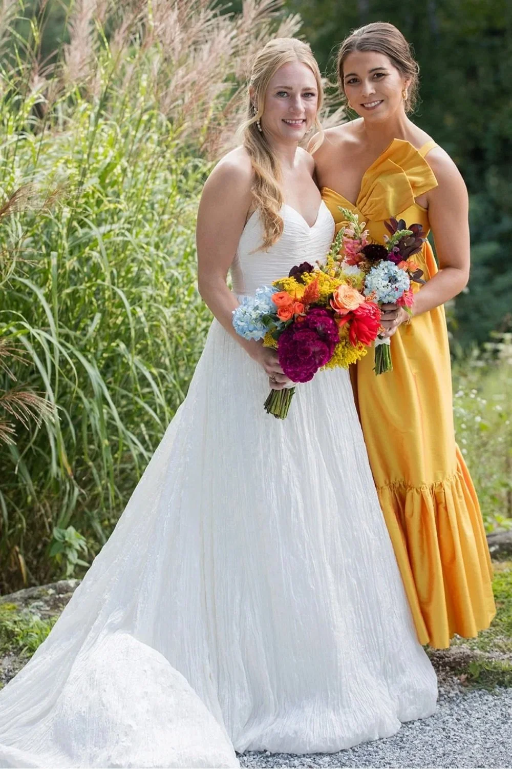 Two women, one in a white wedding dress and the other in a yellow dress, standing outdoors with greenery in the background, holding bouquets of colorful flowers and smiling.