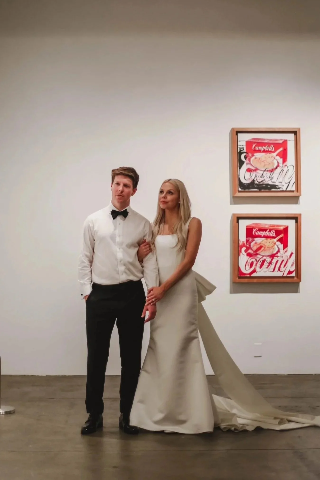A man and a woman dressed in formal attire, standing together in an art gallery. The man is wearing a white shirt with a black bow tie and black pants, while the woman is wearing a white gown. They are standing in front of two framed pop art prints of Campbell's soup cans on the white gallery wall.