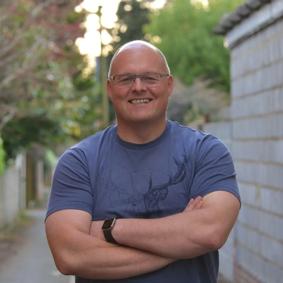 A smiling, bald man with glasses wearing a blue t-shirt and a black watch, standing outdoors with trees and a brick wall in the background.