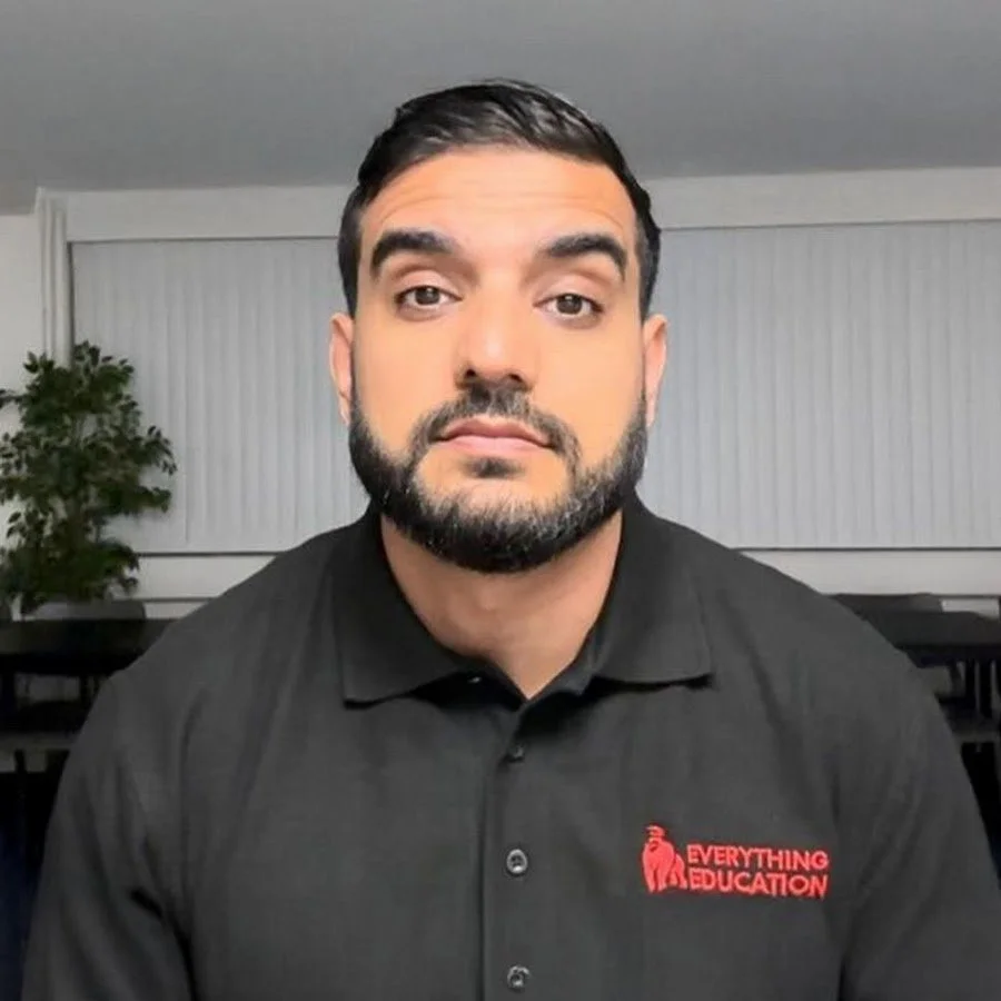 A man with dark hair and a beard wearing a black polo shirt with red text that says "Everything Education". He is sitting indoors with a green plant and white blinds in the background.