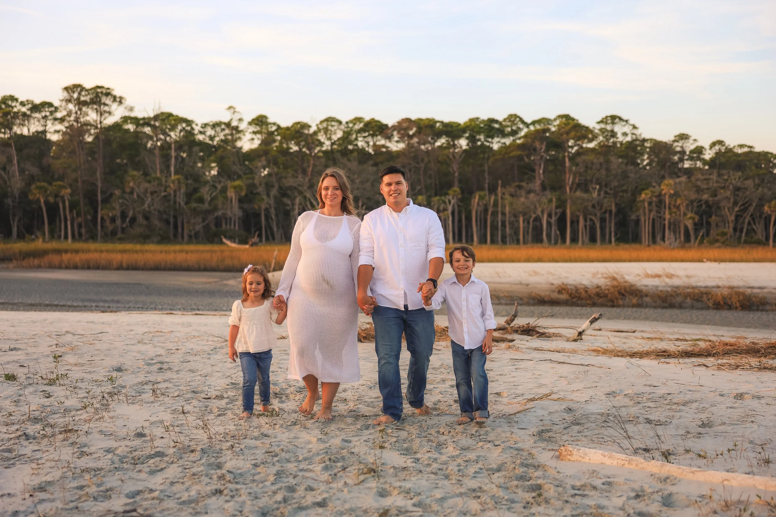 A family of four walking hand in hand on a sandy beach with water and trees in the background during sunset.