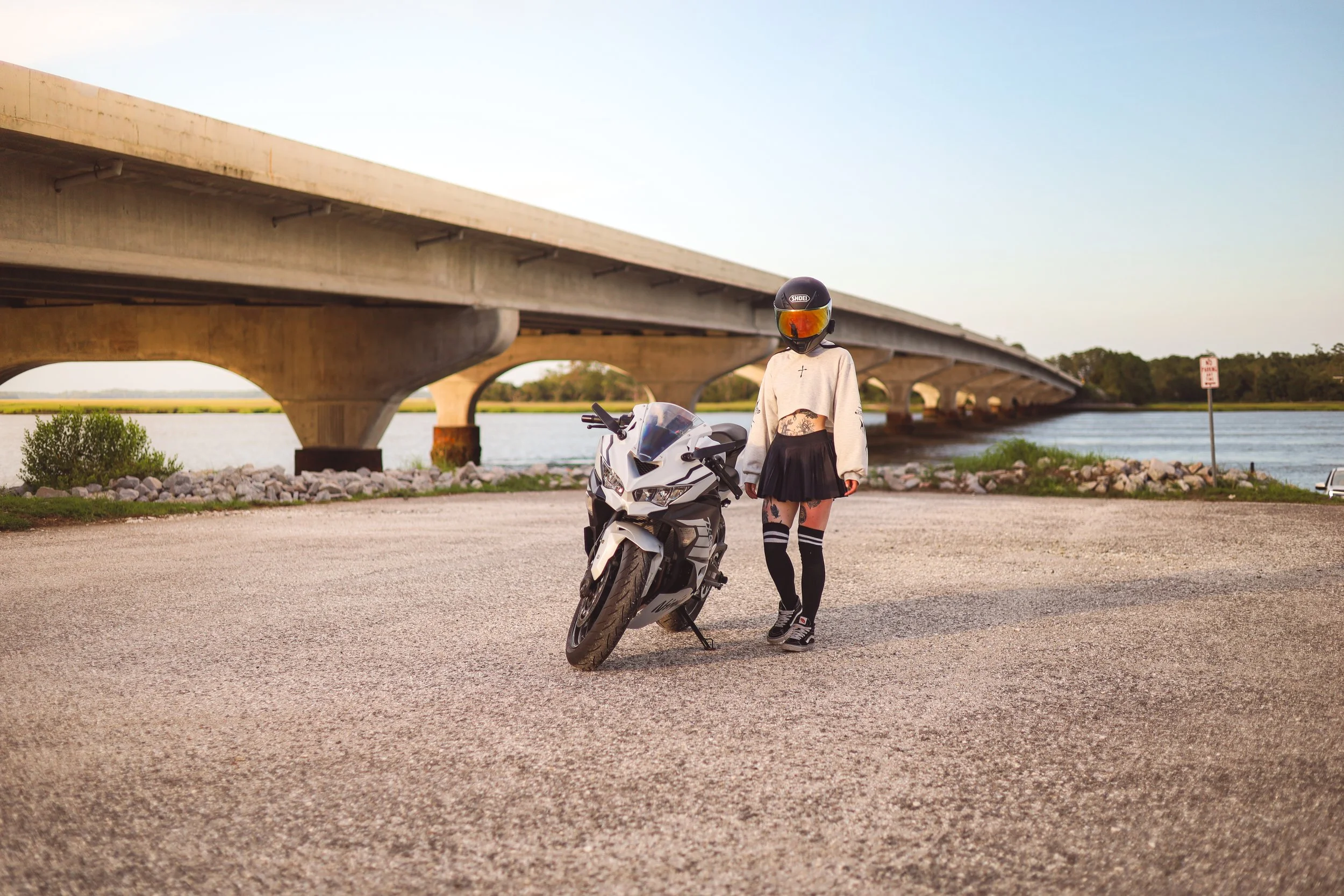 A person in a motorcycle helmet and sporty outfit standing next to a white motorcycle near a bridge over water during sunset.