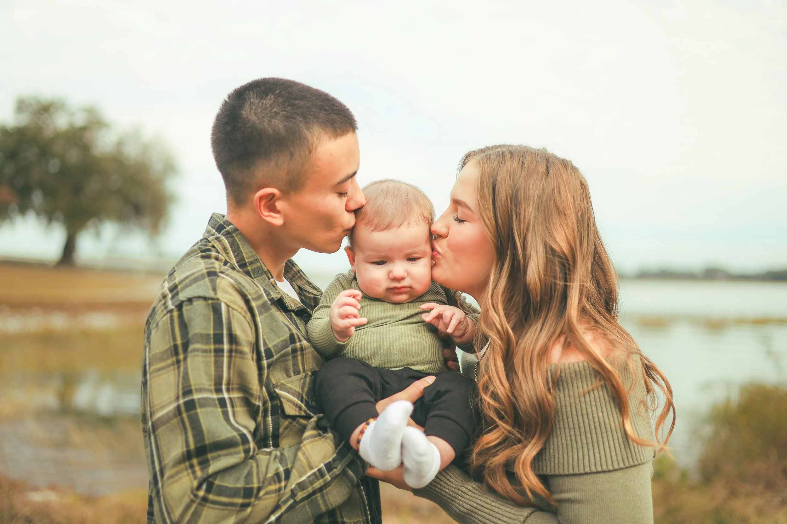 A young couple kissing their baby on the forehead outdoors near a body of water, with trees in the background.