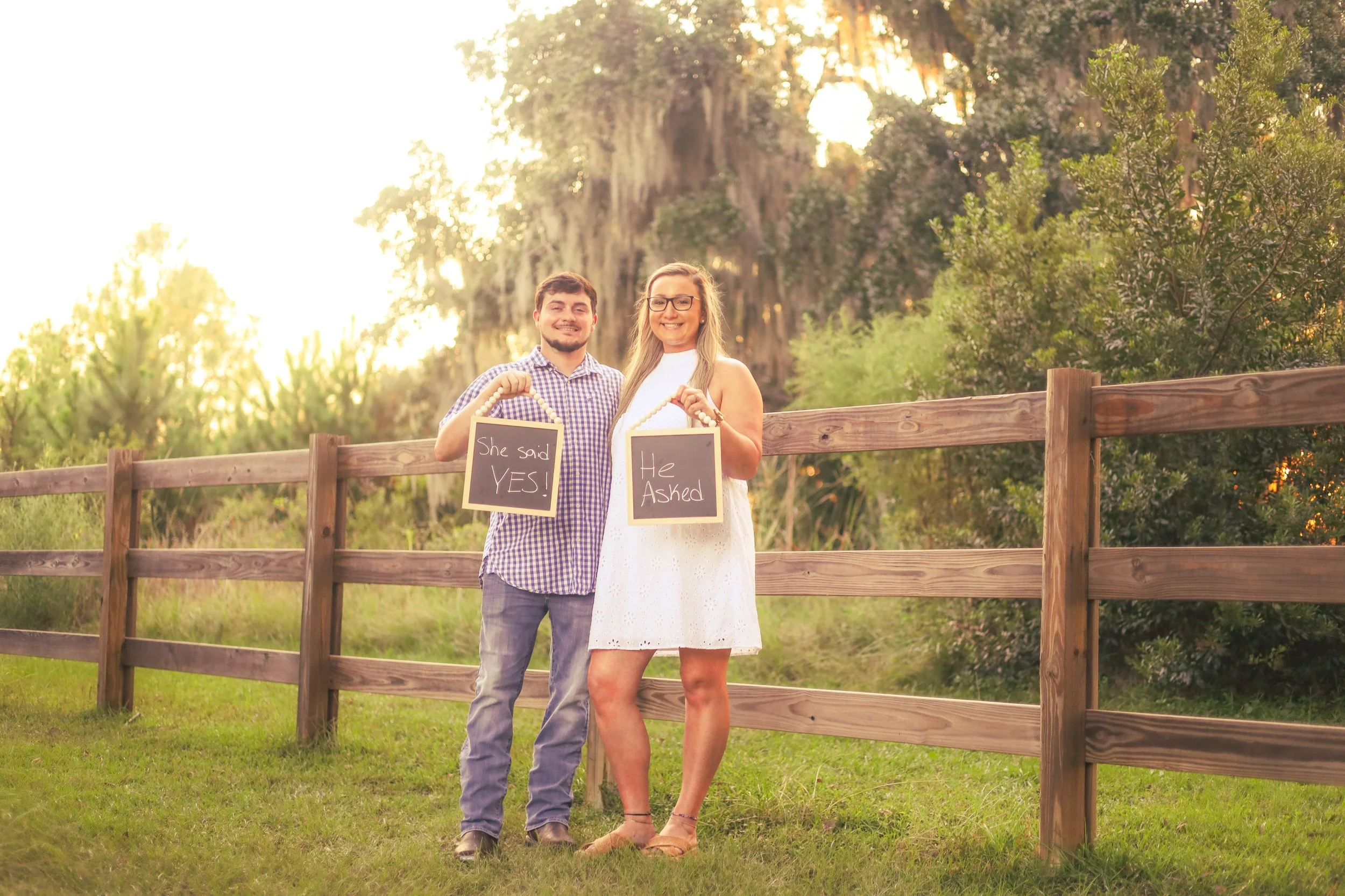A smiling couple standing outdoors by a wooden fence in a sunny, green area, each holding small chalkboard signs with the woman's saying 'She said YES!' and the man's saying 'He Asked'.
