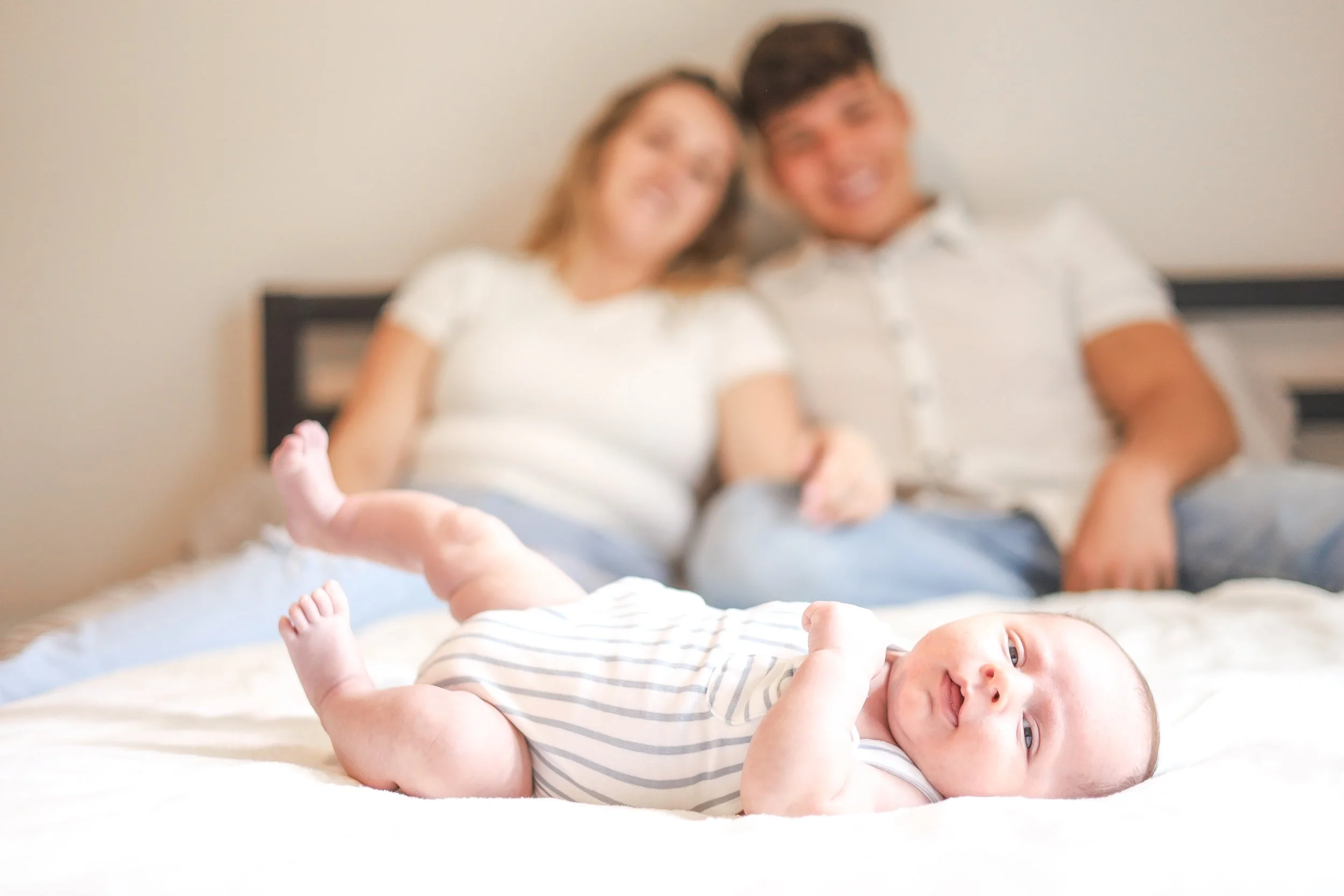 A baby lying on a bed with a smiling expression, in focus, while a couple in the background out of focus look at the camera.