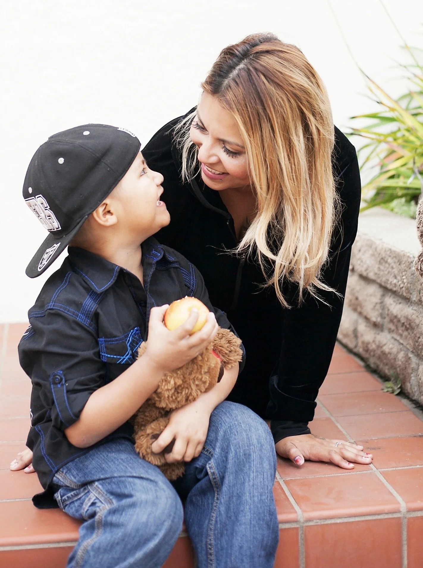 Young child wearing baseball hat poses with teddy bear and mom.
