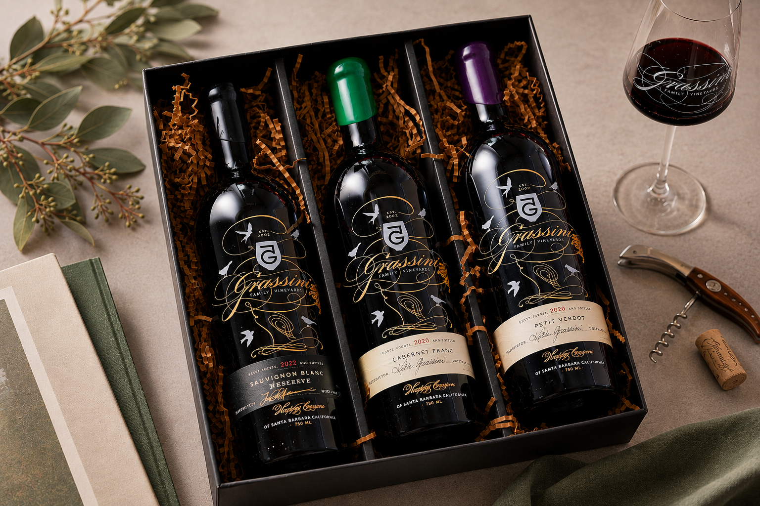 3 bottes of Grassini wine in a black box with gold filler on a table with a glass of wine and florals.