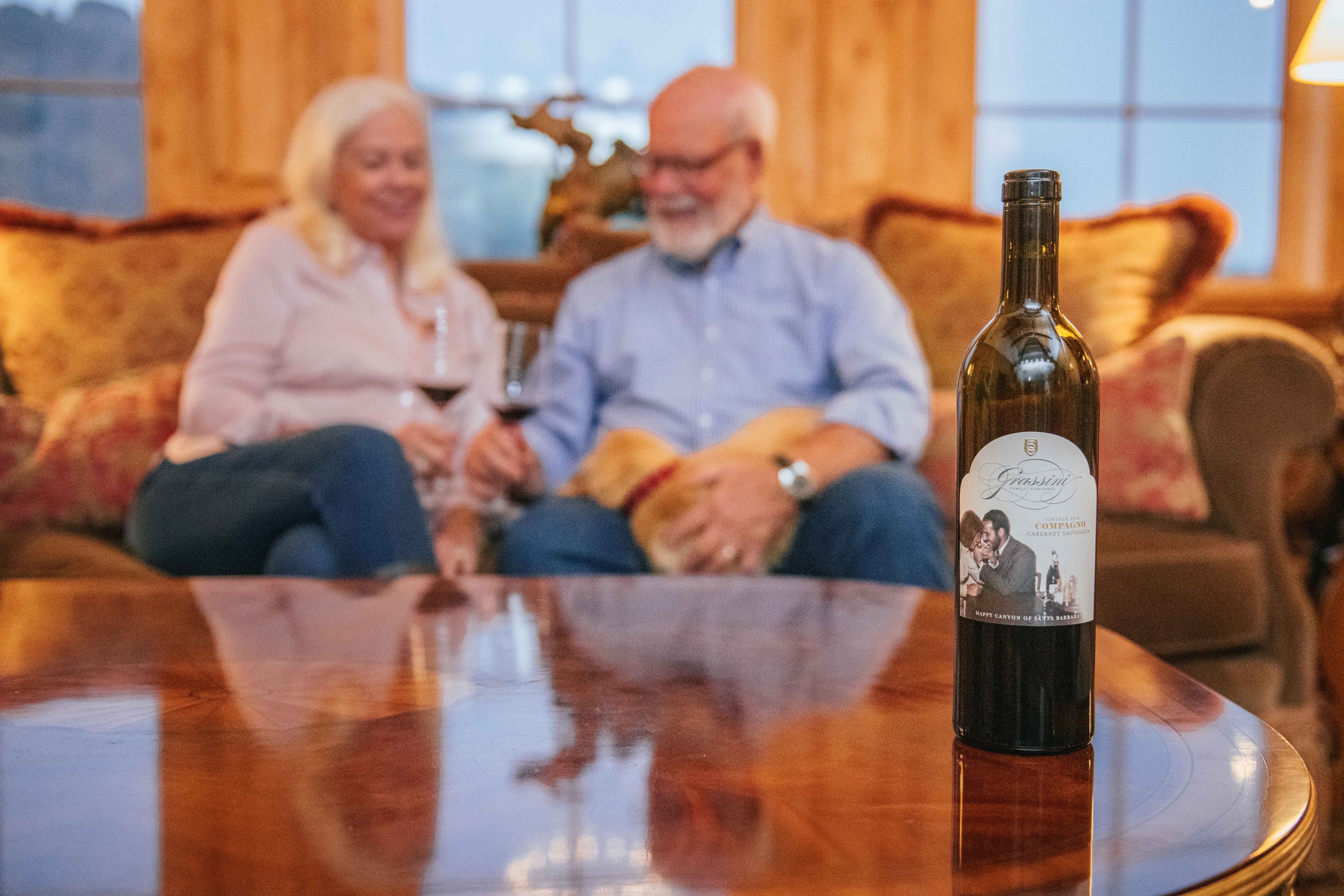 Sharon and Larry Grassini sit on the couch at the winery at the Grassini Family Vineyards Estate with their young pup cuddled up.