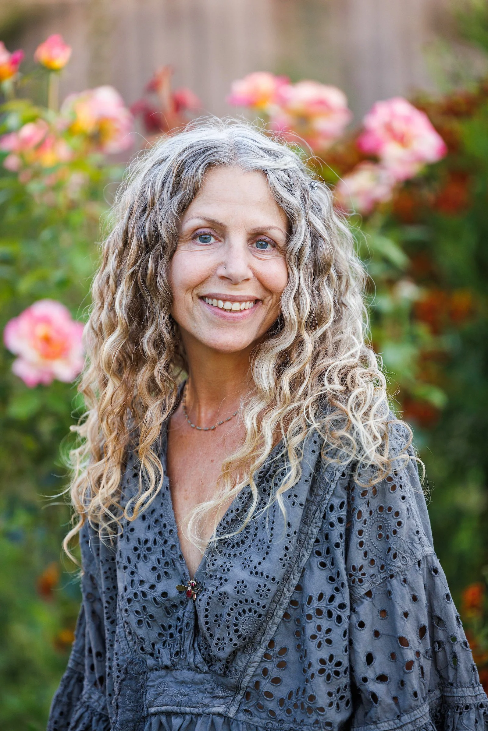 Dina Saalisi author and speaker, a woman with long curly gray hair smiling outdoors with pink flowers in the background.