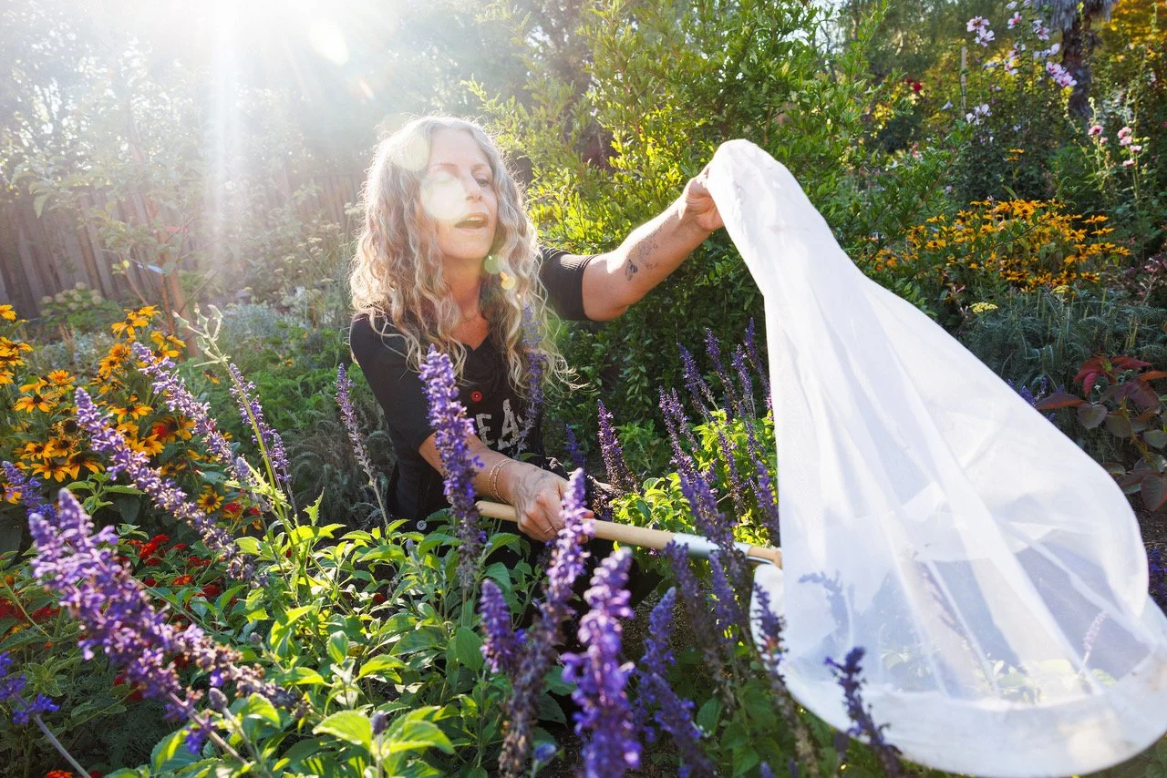 Dina Saalisi, author and speaker, in a garden with colorful flowers, using a white net to gather insects or plants, with sunlight shining through the trees.