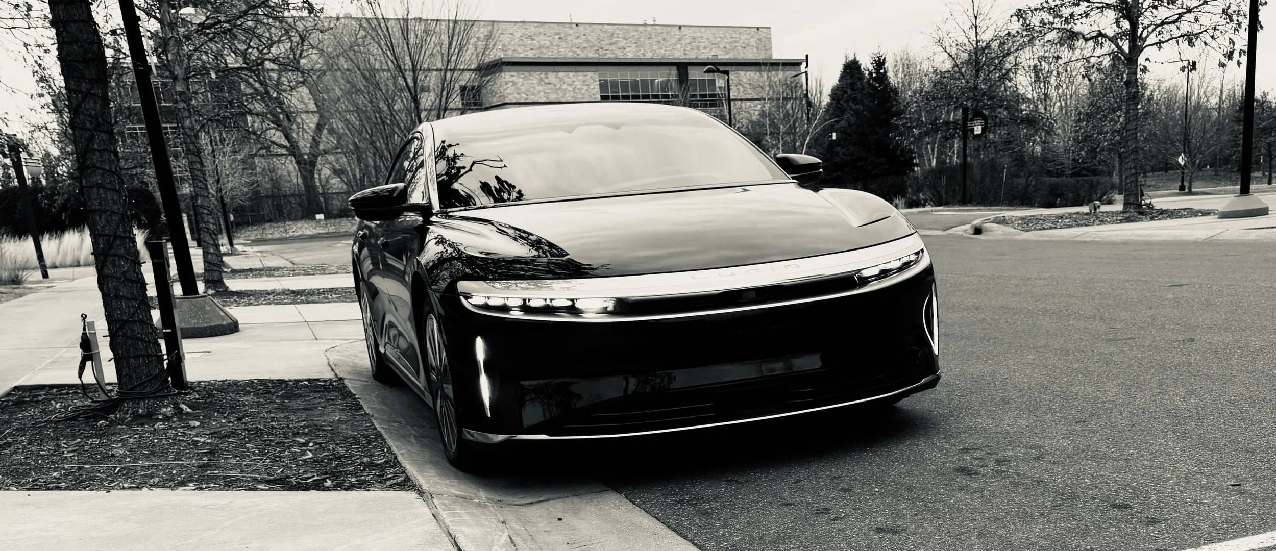 A black Lucid Air electric car parked near a sidewalk on a city street, with trees and a modern building in the background.