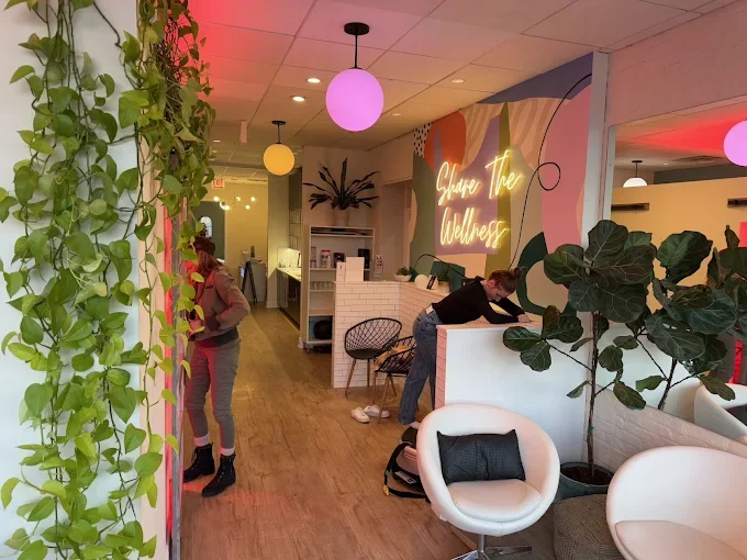 Interior of a wellness center with modern decor, greenery, and a neon sign that says 'Shine the Wellness.' People are preparing or cleaning the reception area.