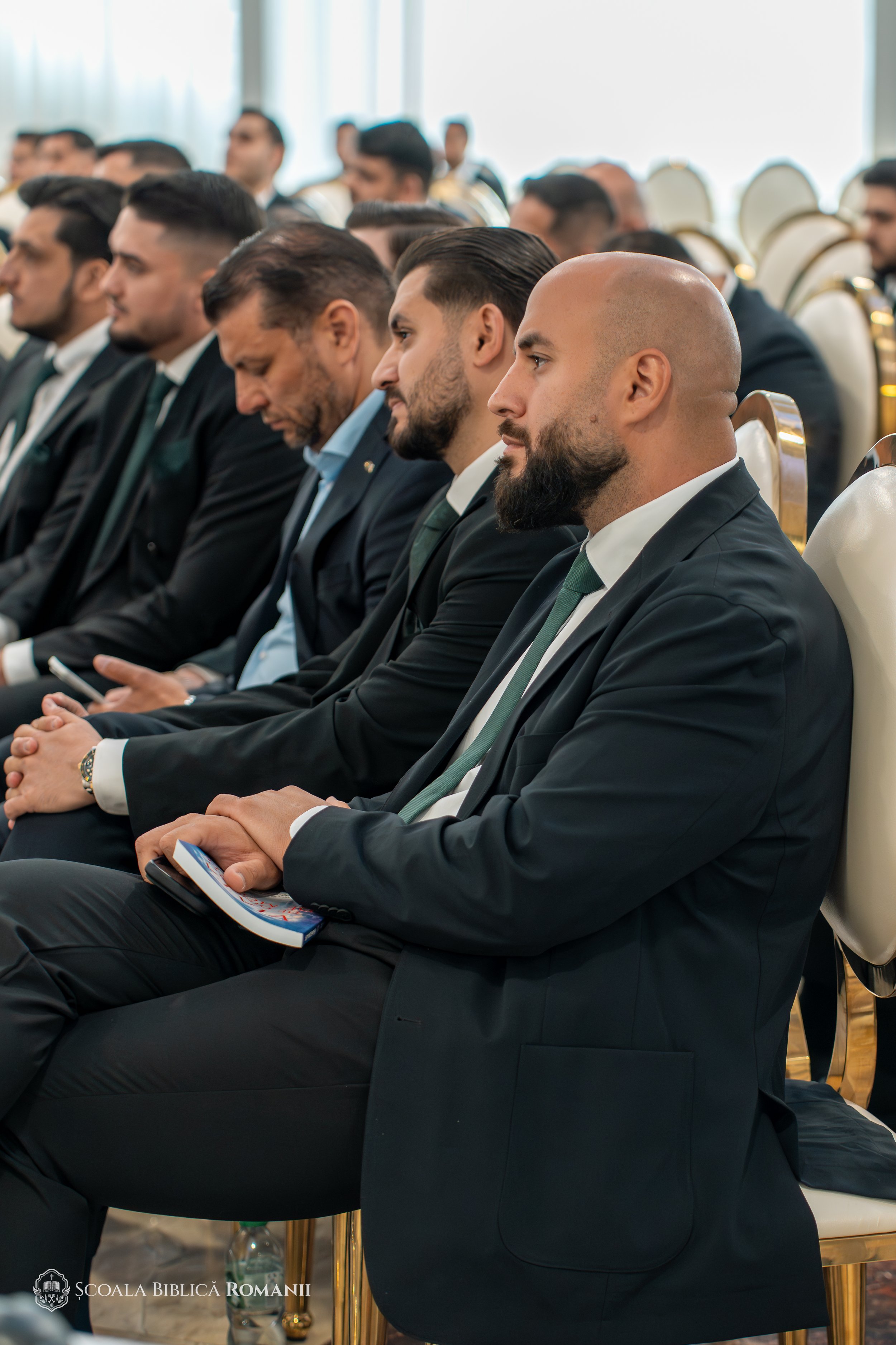 Participanți în sală la ceremonia de absolvire, cu instrumente de alamă în fundal.