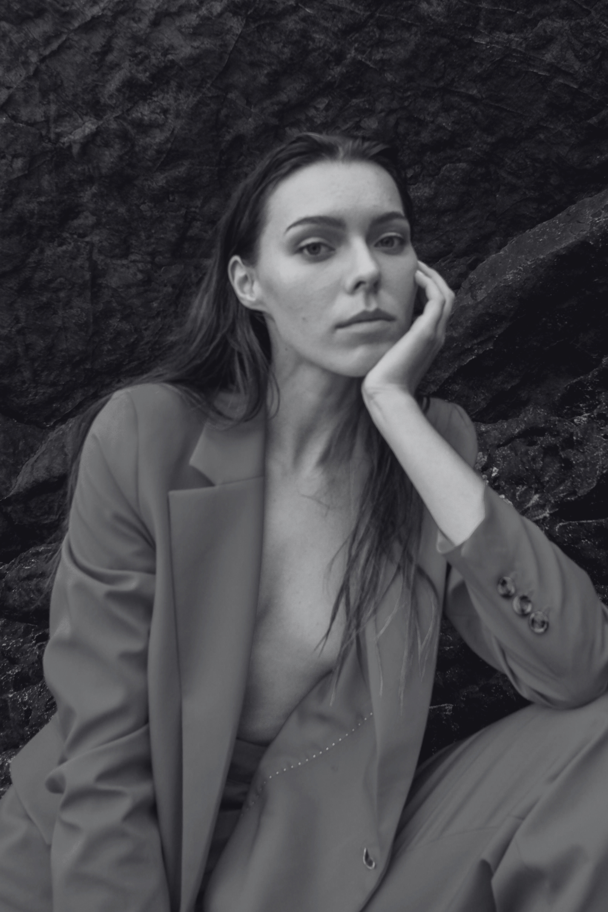 Retrato en blanco y negro de una mujer mirando a la cámara, sentada junto a rocas, con expresión pensativa, sosteniéndose la mejilla con la mano derecha, ropa formal