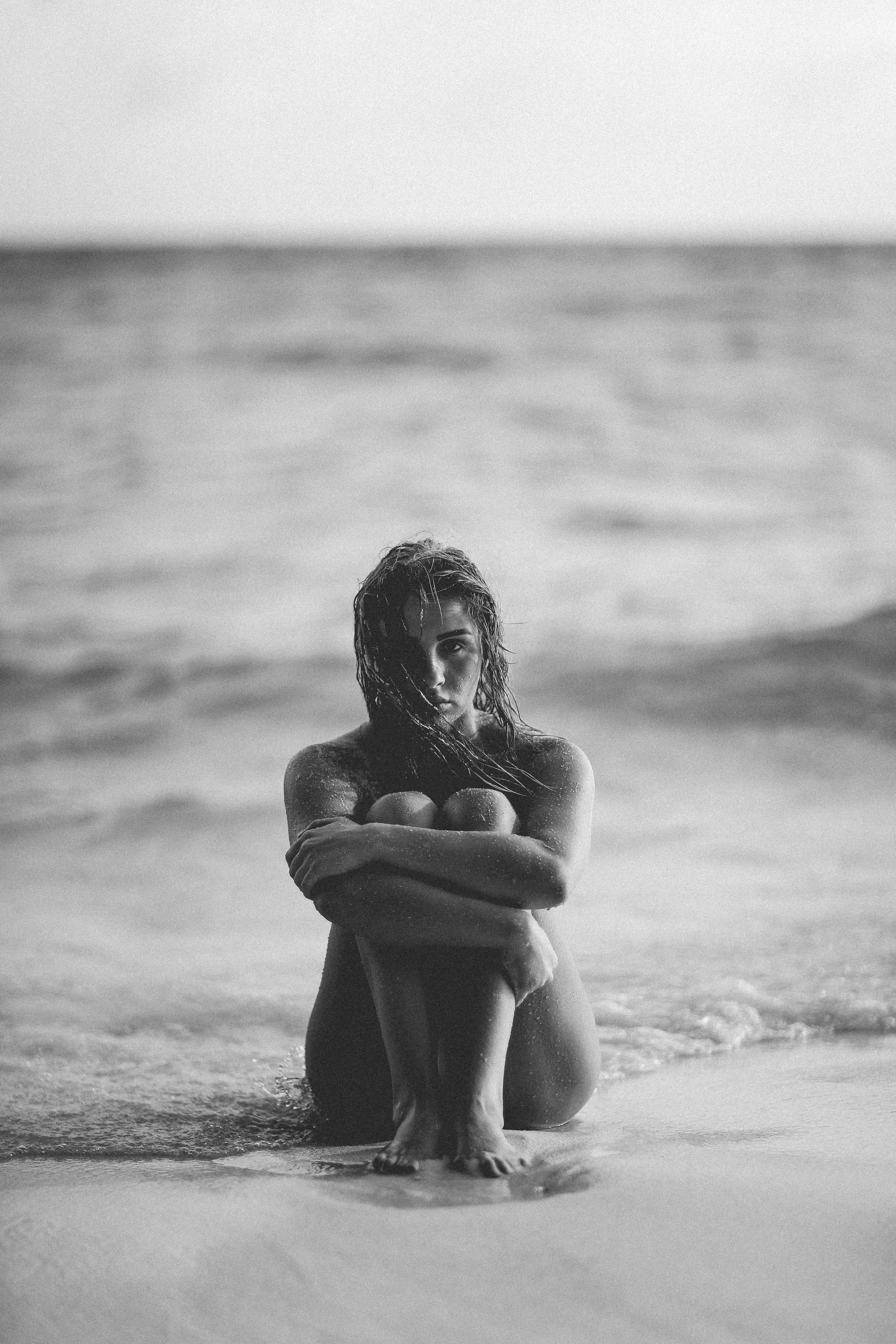 Mujer desnuda en la orilla del mar, en posición de cuclillas, con cabello mojado y mirada fija a la cámara, en fotografía en blanco y negro.