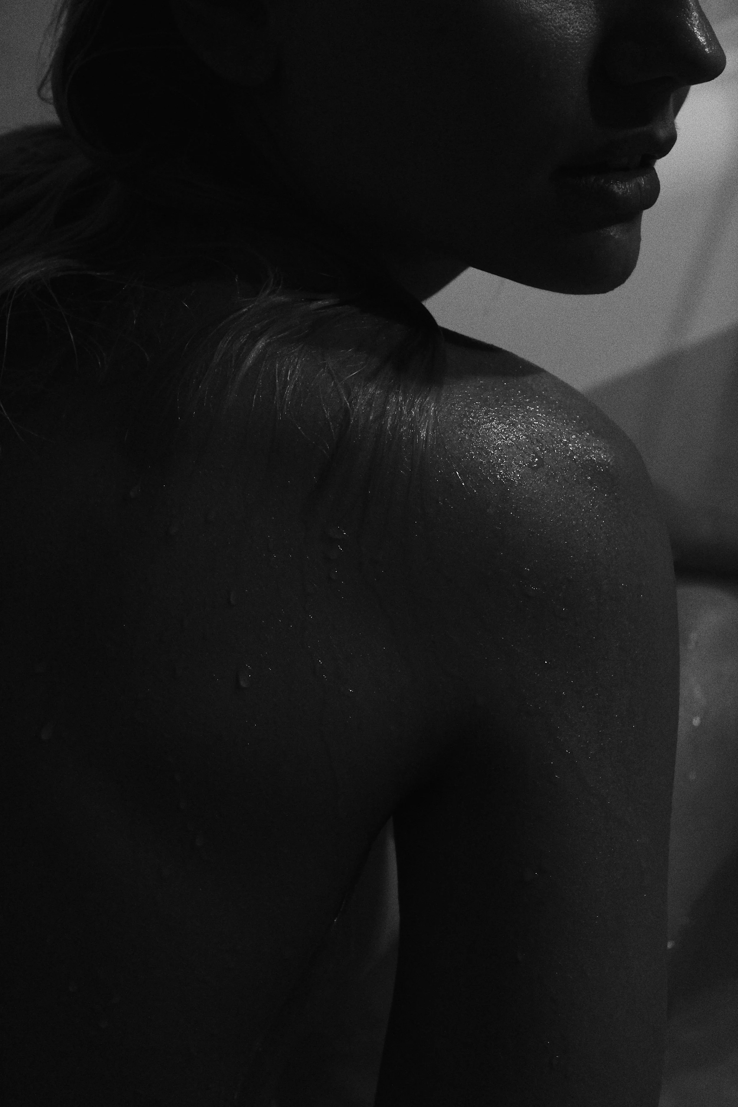Retrato en negro y blanco de una mujer con el rostro parcialmente visible, mostrando su hombro y parte de su cuello, con gotas de agua en su piel.
