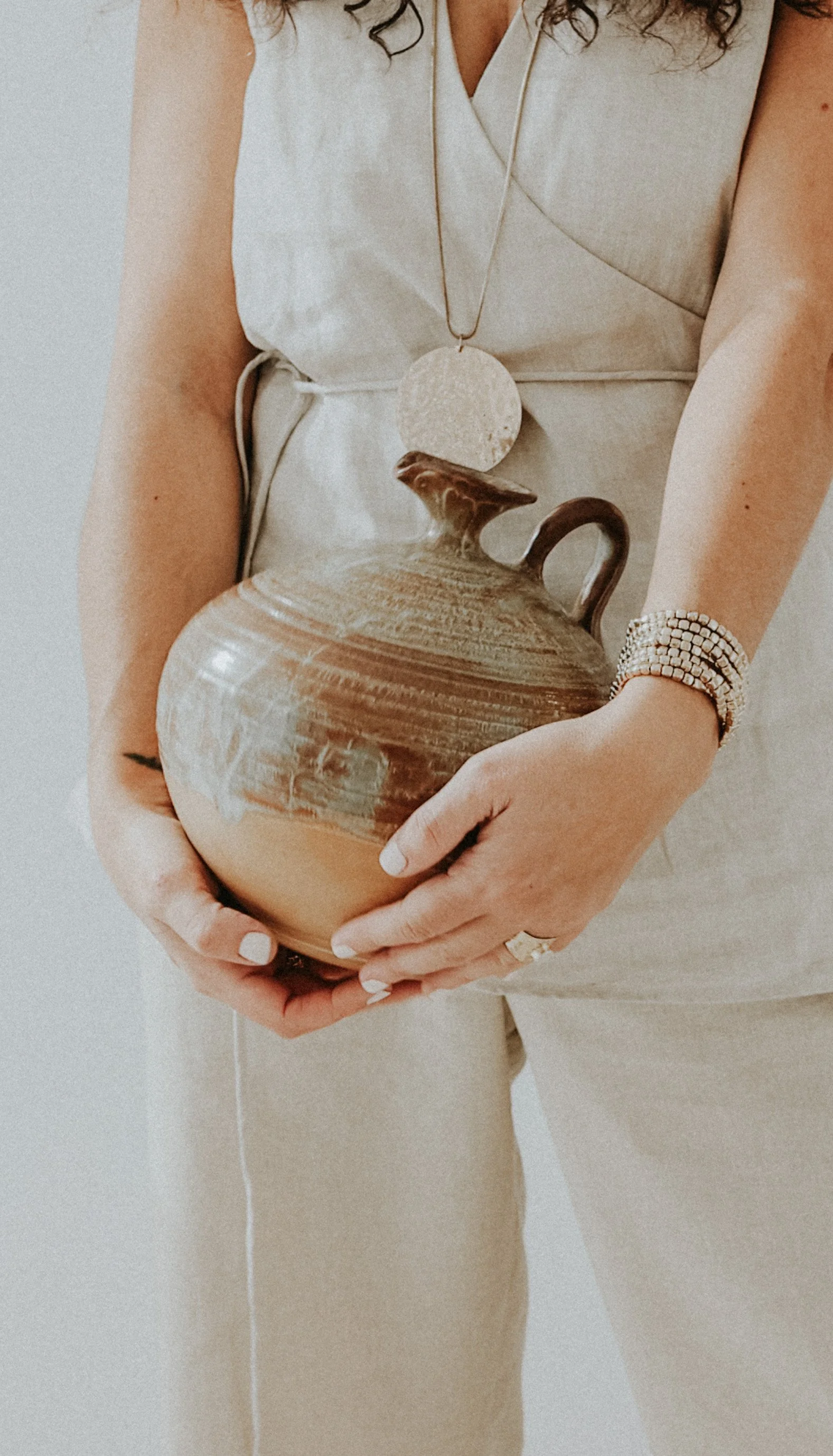Persona sosteniendo una vasija de cerámica con decoración en tonos tierra, usando pulseras y un colgante beige, con ropa blanca y beige.