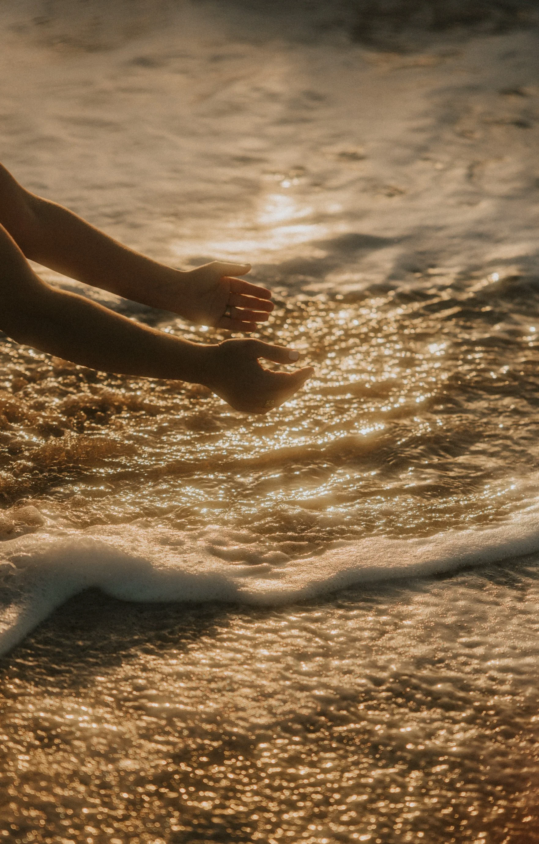 Manos extendidas tocando las olas al atardecer en la playa con reflejos dorados en el agua.