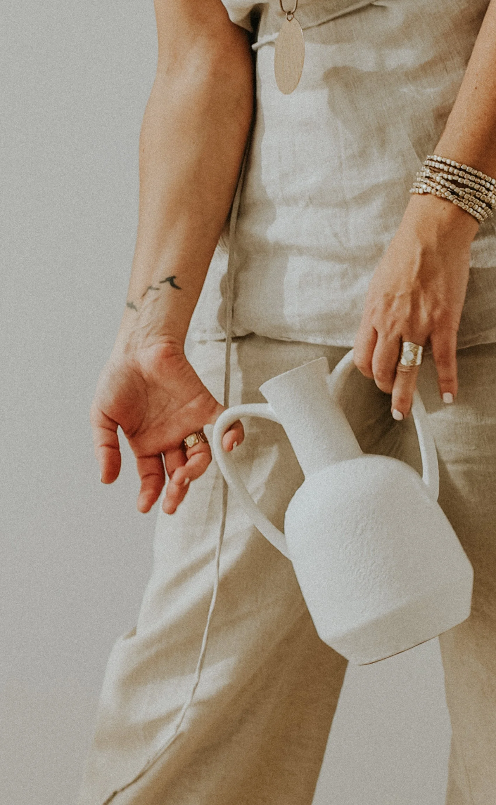 Persona con tatuaje en el brazo, varios anillos, pulseras y un vestido claro, sostiene una jarra blanca de cerámica en un entorno minimalista.