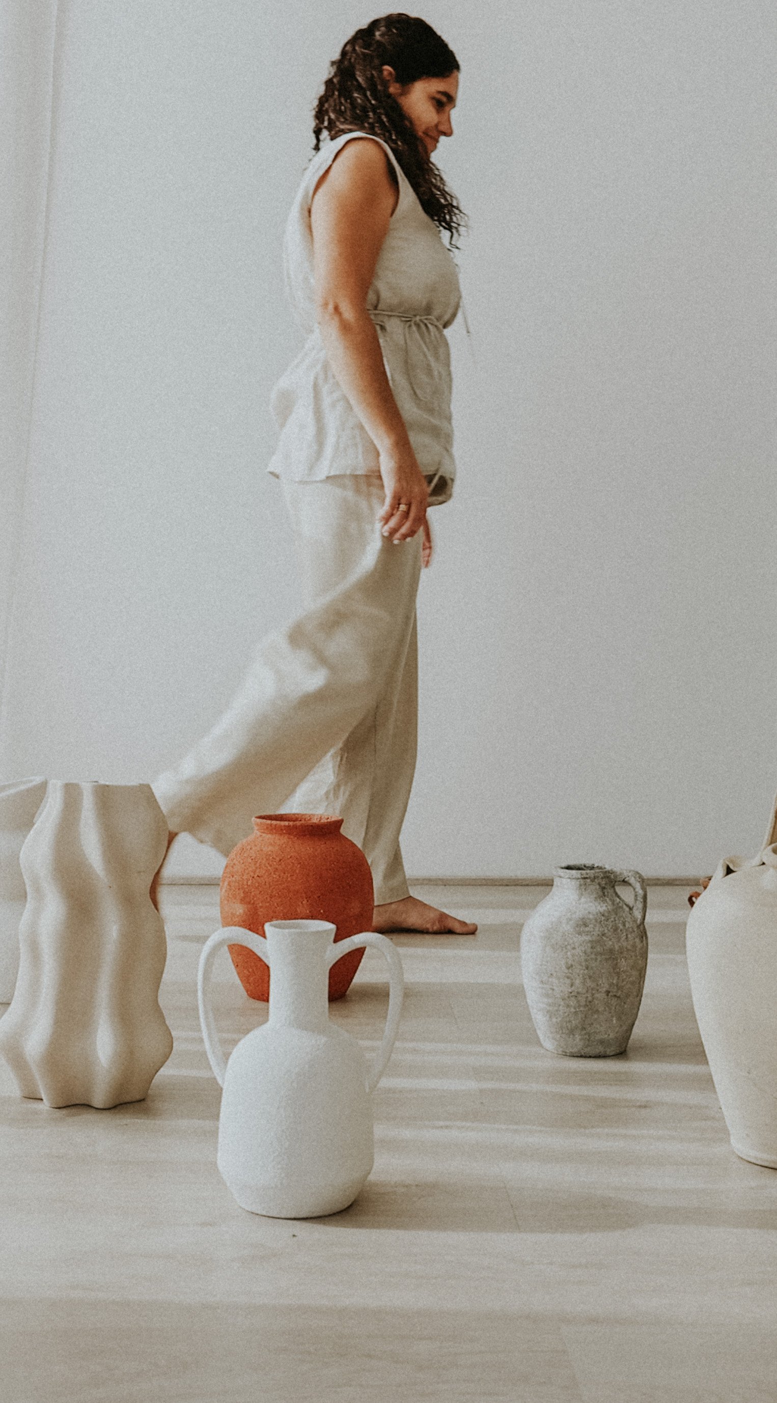 Mujer sonriendo, vestida con ropa clara, rodeada de diferentes vasijas de cerámica de varios tamaños y colores
