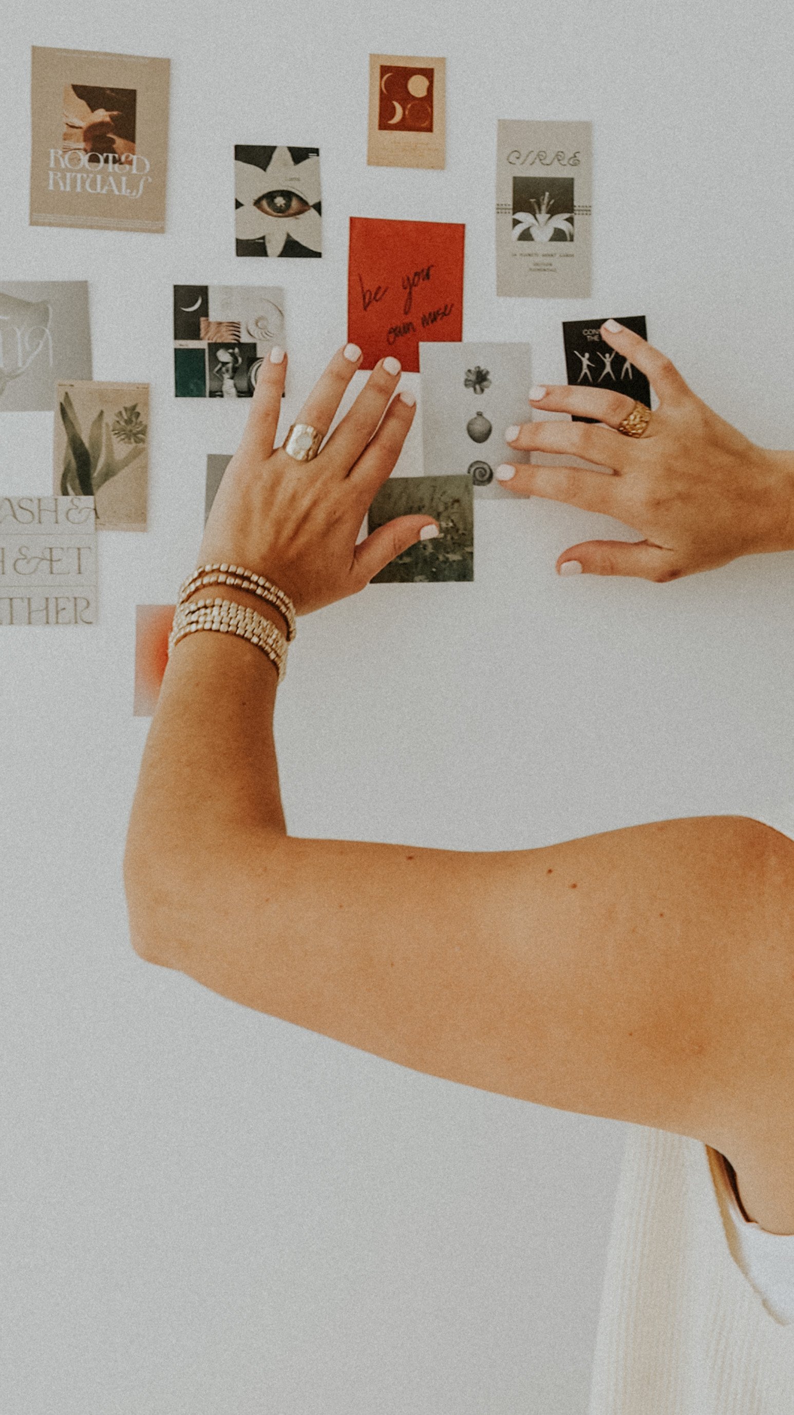 Manos adornadas con pulseras y anillos ajustan notas y recortes en un tablero blanco con varias imágenes y citas, incluyendo una que dice 'be your own muse'.