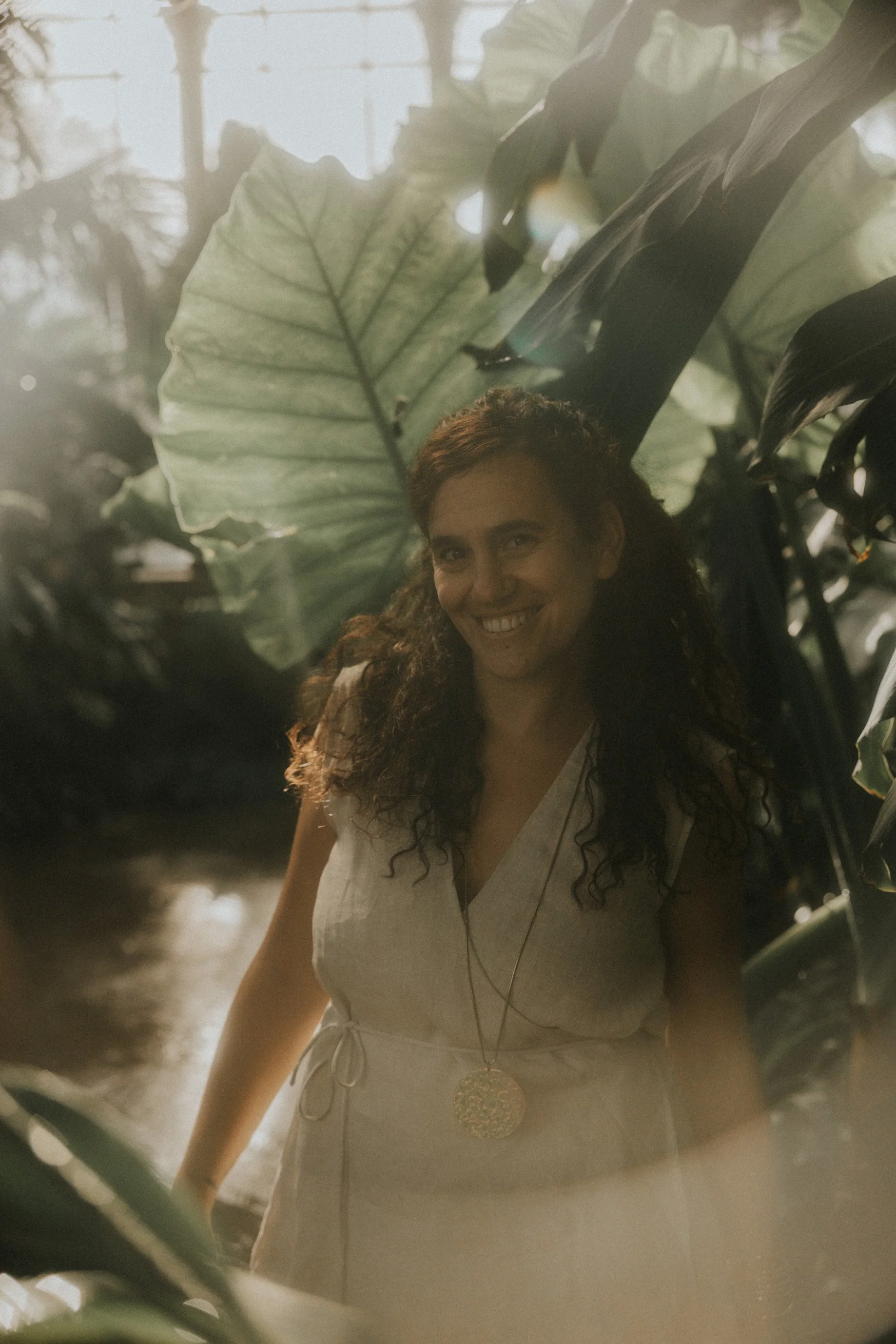 Una mujer con cabello rizado y de sonrisa amplia, vestida con un vestido sin mangas de color claro, posando entre plantas grandes y verdes con luz suave y natural que parece provenir de una ventana o claraboya.