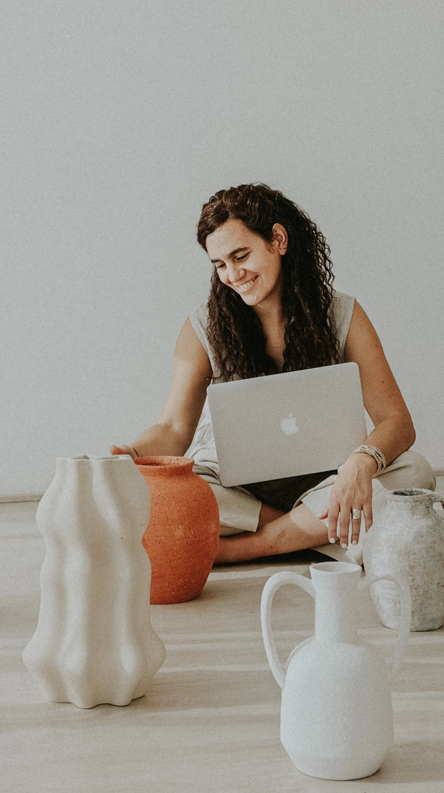 Mujer sonriendo usando una laptop, rodeada de varias jarras de cerámica en un piso de madera.