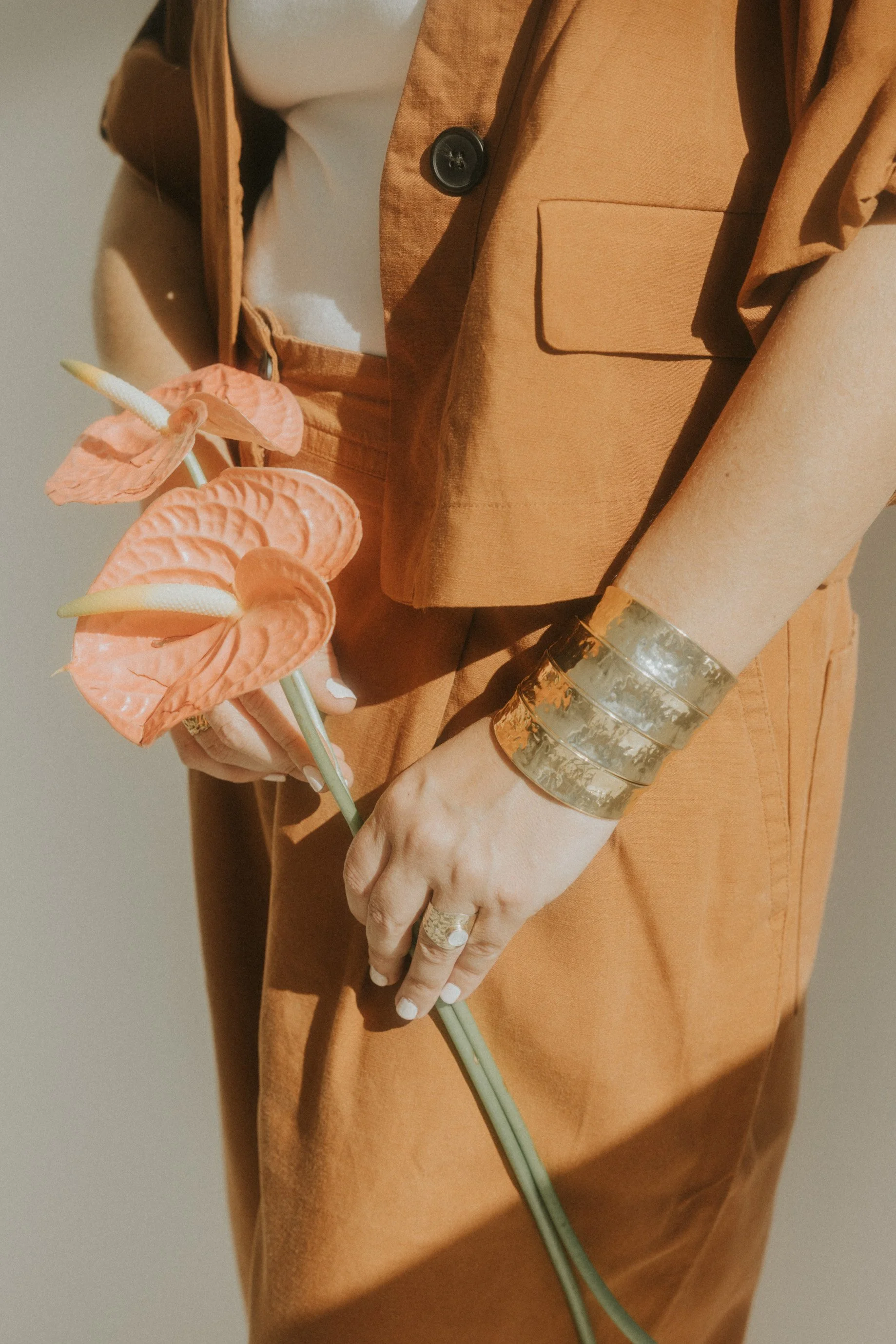 Persona con ropa anaranjada sosteniendo un ramo de flores anthurium de color coral y amarillos, usando pulseras gruesas de metal y anillo.