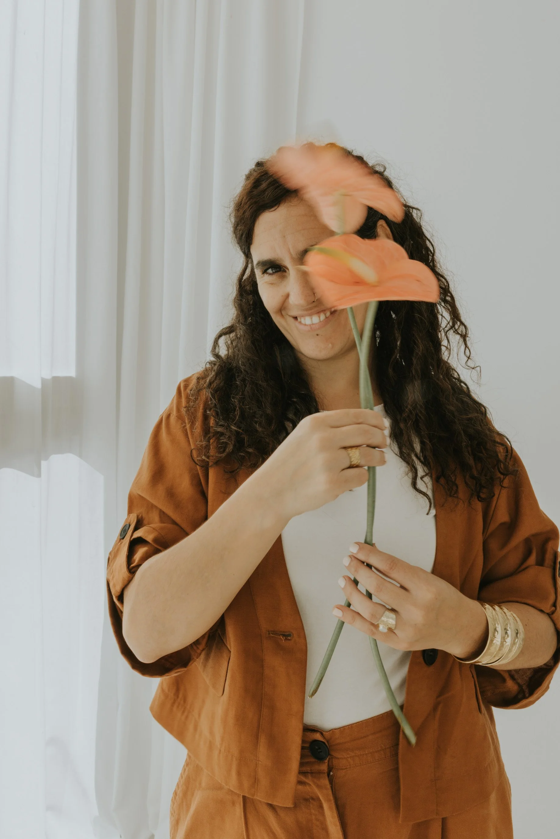 Mujer con cabello rizado, usando ropa de color marrón, sosteniendo un ramo de flores anaranjadas, sonriendo frente a una cortina blanca.