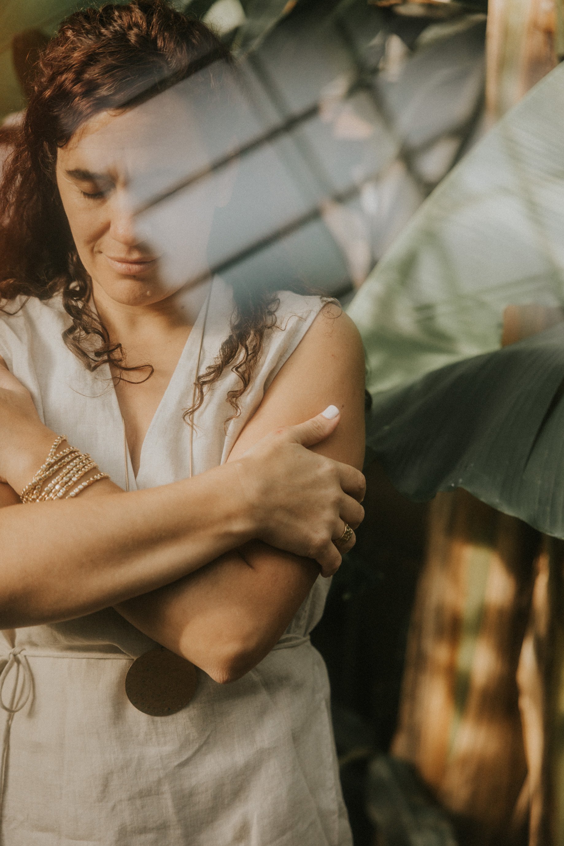 Mujer con cabello rizado y vestido de color crema, con pulseras doradas en la muñeca, abrazándose a sí misma mientras está semitapada por hojas de planta, en un ambiente con luz cálida.