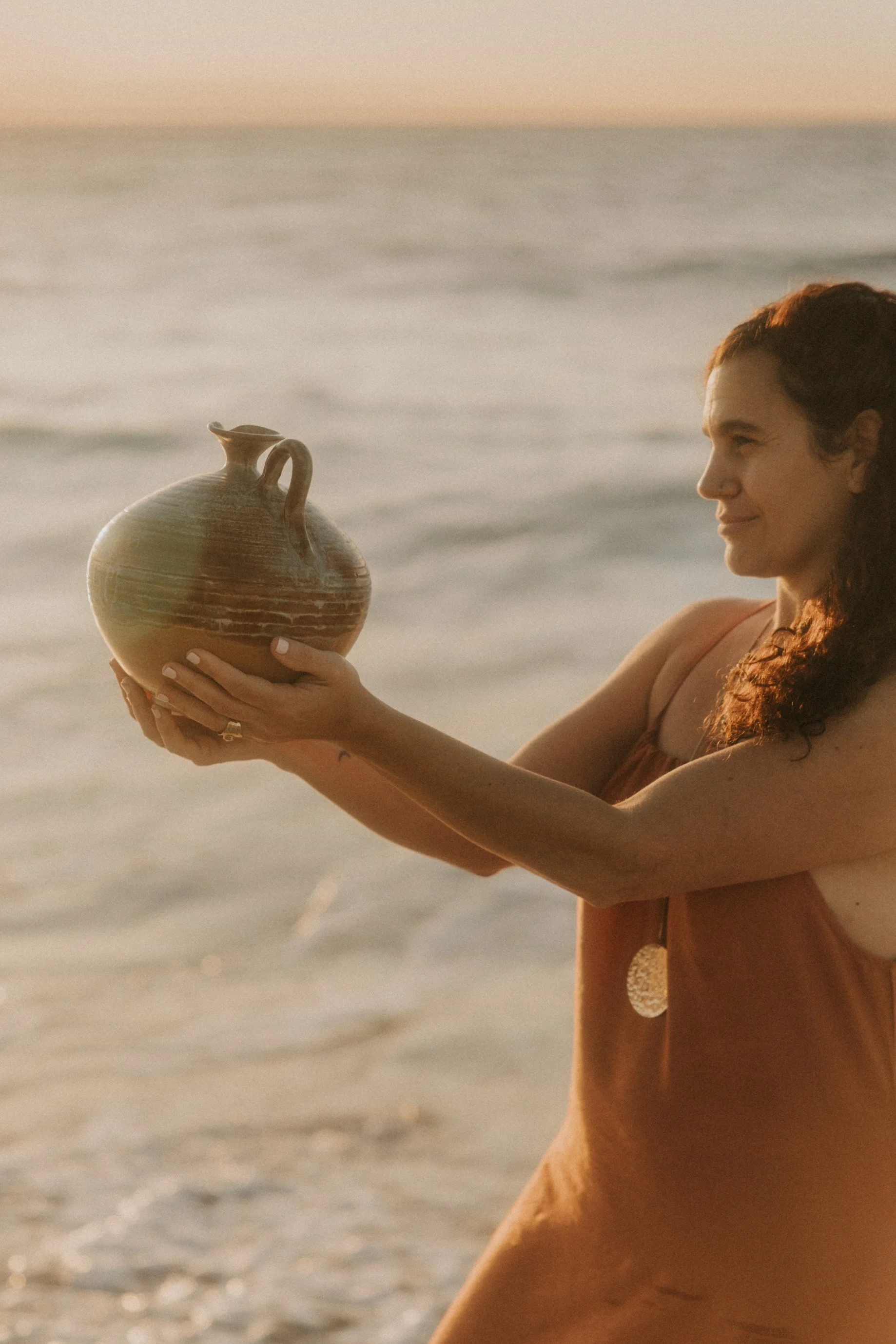 Mujer en la playa sosteniendo una vasija de cerámica durante la puesta del sol.